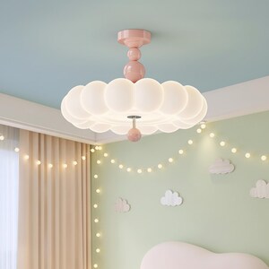 May include: A pink and white ceiling light fixture with a cloud-like design. The light has a pink base and stem, with a series of white, rounded, cloud-shaped lights. The fixture is hanging from a light blue ceiling.