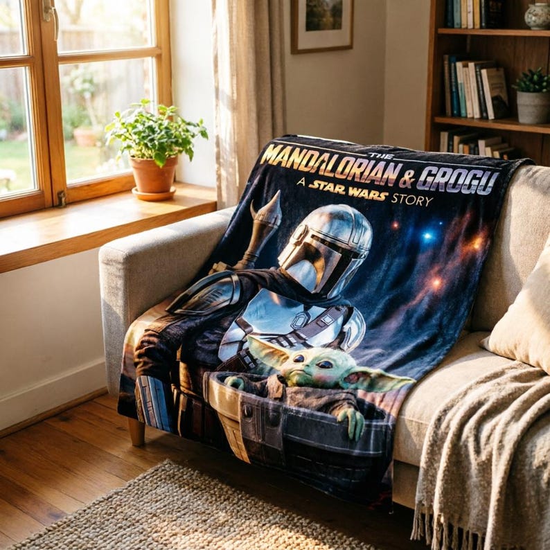 May include: A dark blue throw blanket featuring The Mandalorian and Grogu from A Star Wars Story. The blanket displays the Mandalorian in his armour and Grogu in his carrier. The blanket is draped over a grey sofa.