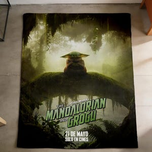 May include: A rectangular blanket featuring a scene from The Mandalorian and Grogu. The image shows Grogu sitting on a mossy log in a forest setting. The blanket has the text "THE STAR WARS MANDALORIAN AND GROGU" and "21 DE MAYO SOLO EN CINES."