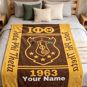 Op de afbeelding: Een bruine en gouden deken met de Griekse letters Iota Phi Theta en het jaar 1963. De deken heeft een wapen met sterren en een schildontwerp. De woorden "Iota Phi Theta" staan op de zijkanten en "Your Name" onderaan.