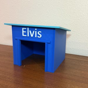 May include: A blue pet house with a light blue slanted roof. The front of the structure features the name Elvis in white, raised lettering above a rectangular opening. The design includes a frame around the entrance and solid blue side walls.