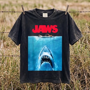 May include: A black short-sleeve t-shirt featuring the Jaws movie poster graphic. The design displays the red text JAWS above an illustration of a swimmer on the surface of the water with a large shark swimming upward from the depths below.
