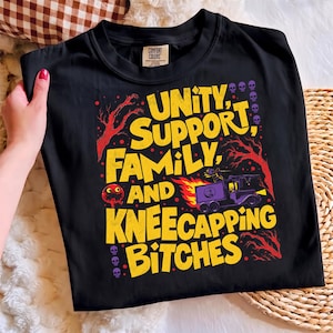 May include: A black t-shirt featuring a graphic print with yellow text that reads "UNITY, SUPPORT, FAMILY, AND KNEECAPPING BITCHES." The design includes red tree silhouettes, a purple truck with flames, a small red circular character, and purple skulls.