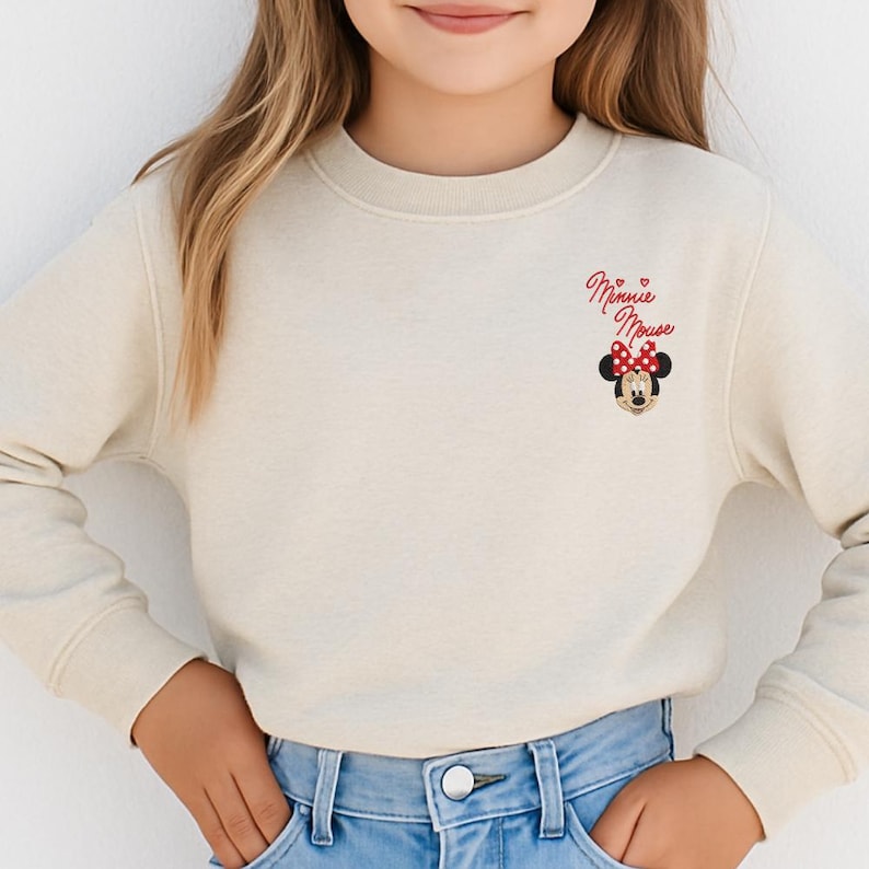 A young girl wearing a white sweater with a Minnie Mouse design and a red bow on her head.