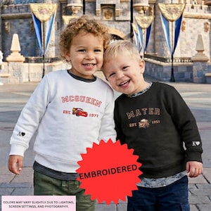 Two young children, a boy and a girl, are standing together and smiling in front of a castle. They are wearing sweatshirts with the text "McQueen" and "Mater" printed on them.