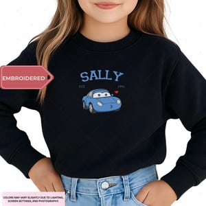 A young girl wearing a black sweater with a blue car embroidered on it.