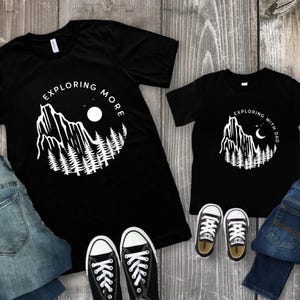 May include: Black t-shirts with white graphic designs. One shirt says "EXPLORING MORE" and the other says "EXPLORING WITH DAD". The designs feature a mountain, trees, and a moon. Black sneakers and blue jeans are also visible.