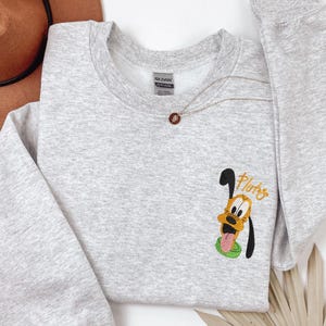 A gray sweatshirt with a cartoon character printed on it, specifically a dog. The character is wearing a green hat and has a tongue sticking out. The sweatshirt also has a small embroidered patch on the front that says "Pluto".