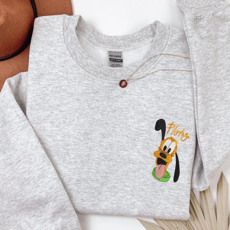 A gray sweatshirt with a cartoon character printed on it, specifically a dog. The character is wearing a green hat and has a tongue sticking out. The sweatshirt also has a small embroidered patch on the front that says "Pluto".