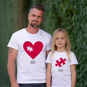 May include: White t-shirts with matching puzzle piece designs. One shirt features a red heart with a puzzle piece and the word "Daddy." The other shirt has a red puzzle piece and the name "Sophia."