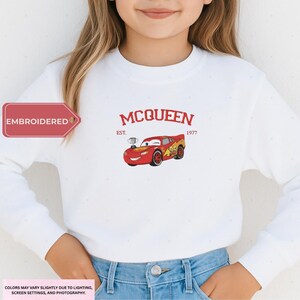 A young girl wearing a white sweatshirt with a red and yellow car design and the text "McQueen" printed on it.
