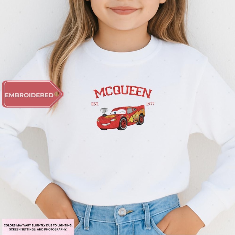 A young girl wearing a white sweatshirt with a red and yellow car design and the text "McQueen" printed on it.