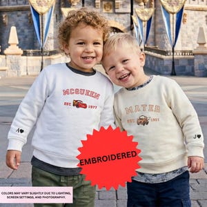 Two young boys wearing matching sweatshirts with the character "Mate" on them, smiling and posing together in front of a castle-like structure.