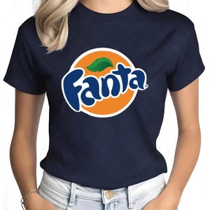 May include: Navy blue t-shirt featuring the Fanta logo. The logo is a white-outlined blue wordmark over an orange circle, with a green leaf accent. The shirt is a classic crew neck style.