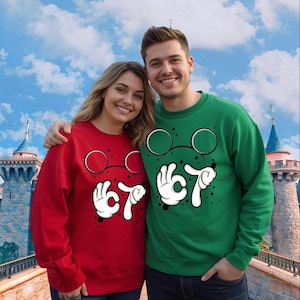 A smiling couple wearing matching red and green sweaters with cartoon-style glasses and hand gestures stands in front of a castle-like structure.