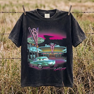 Può includere: Una t-shirt nera a maniche corte con una stampa grafica di un'auto color verde acqua davanti al Flo's V8 Cafe. Il design include luci al neon rosa e blu, il testo Flo's V8 Cafe e la scritta Radiator Springs nella parte inferiore.