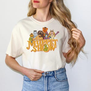 Puede incluir: Una camiseta de manga corta de color crema con un estampado gráfico de varios personajes de los Muppets, incluyendo a la rana Gustavo, la cerdita Peggy y el oso Fozzie, con el texto The Muppet Show en una fuente naranja de estilo retro.