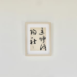 May include: A framed artwork featuring black calligraphy on a cream-colored background. The calligraphy includes four vertical columns of Japanese characters. The frame is a light wood color, and the artwork is displayed against a white wall.