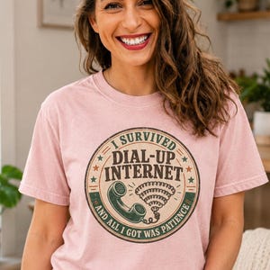 Może przedstawiać: Jasnoróżowy T-shirt z krótkim rękawem z okrągłą grafiką zawierającą napis I SURVIVED DIAL-UP INTERNET AND ALL I GOT WAS PATIENCE oraz ilustrację zabytkowej słuchawki telefonicznej i ikony sygnału.
