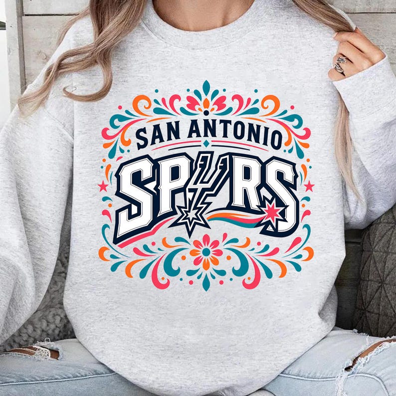 以下が含まれることがあります： 「SAN ANTONIO SPURS」の文字が大胆で様式化されたフォントで描かれた、ライトグレーのスウェットシャツ。デザインには星と、ティール、オレンジ、ピンクの装飾的なフローラル要素が含まれています。カジュアルなアパレルアイテムです。