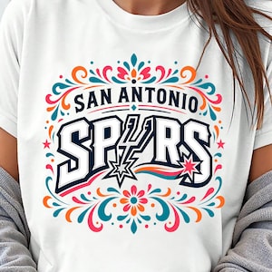 以下が含まれることがあります： 大胆で様式化されたフォントで「SAN ANTONIO SPURS」という文字がプリントされた白いTシャツ。デザインには星と、青、オレンジ、ピンク、ティール色のカラフルな花のアクセントが含まれています。