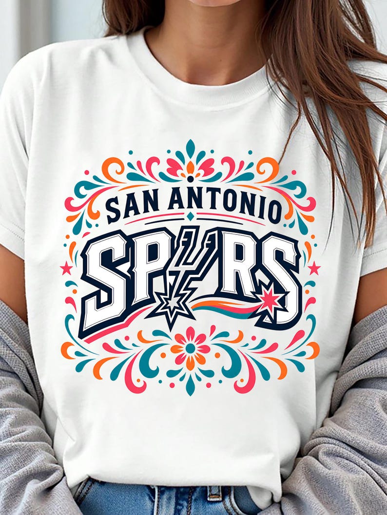 以下が含まれることがあります： 大胆で様式化されたフォントで「SAN ANTONIO SPURS」という文字がプリントされた白いTシャツ。デザインには星と、青、オレンジ、ピンク、ティール色のカラフルな花のアクセントが含まれています。