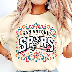 以下が含まれることがあります： 大胆な書体で「SAN ANTONIO SPURS」と書かれたクリーム色のTシャツ。デザインには星と、ターコイズ、オレンジ、ピンクの装飾的な花の要素が含まれています。鮮やかでカラフルなグラフィックTシャツ。