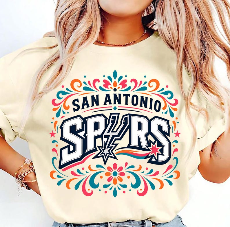 以下が含まれることがあります： 大胆な書体で「SAN ANTONIO SPURS」と書かれたクリーム色のTシャツ。デザインには星と、ターコイズ、オレンジ、ピンクの装飾的な花の要素が含まれています。鮮やかでカラフルなグラフィックTシャツ。