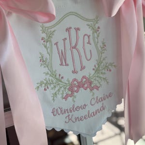 May include: A white fabric banner featuring a pink embroidered monogram WKC inside a light green floral wreath. Below the wreath, the name Winslow Claire Kneeland is embroidered in pink script. The banner is accented with large pink satin bows.