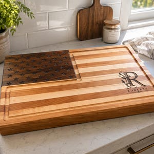 May include: A rectangular wooden cutting board featuring a dark wood section with carved stars and a light wood section with horizontal stripes, resembling an American flag. The board includes a juice groove and the engraved letter R and Roberts.