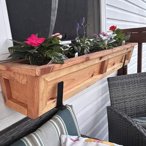 May include: A wooden window box filled with vibrant flowers in shades of pink, purple, and red. The rectangular planter is made of light-colored wood and is mounted on a white exterior wall with black metal brackets.