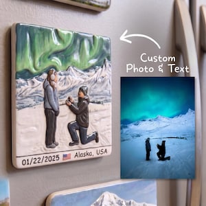 May include: A square ceramic magnet featuring a raised relief scene of a proposal in a snowy landscape under green aurora borealis lights. The text 01/22/2025, a small American flag icon, and Alaska, USA are printed at the bottom of the magnet.