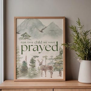 Può includere: Una stampa artistica incorniciata con un'illustrazione ad acquerello di una scena di foresta, tra cui montagne, alberi e un cervo. Il testo "FOR THIS CHILD WE HAVE PRAYED" è in verde, con il versetto biblico 1 Samuele 1:27. La cornice è in legno marrone chiaro.