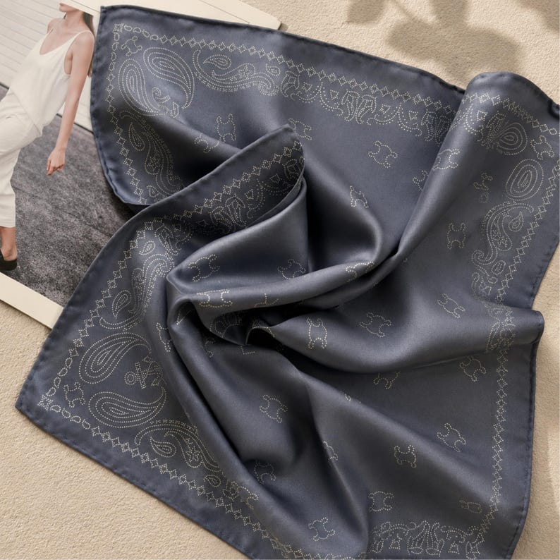 May include: A folded, slate-blue silk scarf with a paisley and bear pattern. The scarf has a smooth, shiny texture and a decorative border. The design is in a lighter shade of blue, creating a subtle contrast.