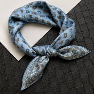 Peut inclure: Un foulard en soie bleu clair orné d'un motif cachemire dans des tons de bleu, marron et blanc. Le foulard est noué, mettant en valeur le design détaillé et la texture du tissu. Le foulard est de forme carrée.