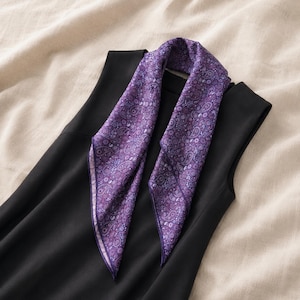 May include: A square purple scarf featuring a detailed paisley pattern in shades of violet, lavender, and light blue. The scarf is draped over a sleeveless black top, showcasing its intricate floral-inspired motifs and finished edges.