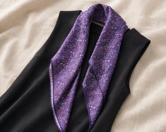 Puede incluir: Un pañuelo cuadrado de color morado con un patrón de cachemira detallado en tonos violeta, lavanda y azul claro. El pañuelo está colocado sobre una blusa negra sin mangas, mostrando sus complejos motivos florales y bordes acabados.