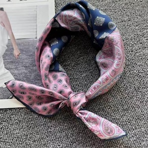 Op de afbeelding: Een geknoopte zijden sjaal met een paisley patroon in roze en marineblauw. De sjaal is in een knoop gelegd, met de uiteinden naar beneden gericht. De sjaal is gemaakt van een zacht, zijdeachtig materiaal.