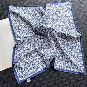May include: A square pocket square featuring a dark blue background with a repeating pattern of small white flowers. The fabric has a solid dark blue border along all four edges and a small white rectangular care label attached to one corner.