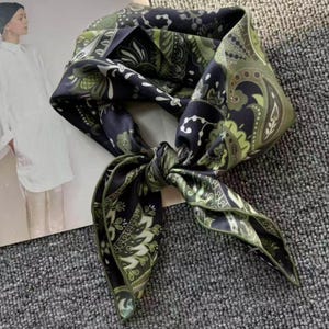 May include: A square scarf featuring a dark navy blue and olive green paisley pattern with intricate white floral and geometric details. The fabric has a smooth, satin-like finish and is tied in a knot, displaying the repeating ornate design.