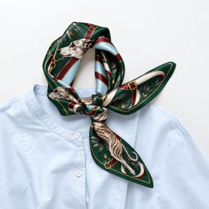 Puede incluir: Un pañuelo de seda verde oscuro con un patrón repetitivo de galgos, hebillas ecuestres doradas y rayas azul claro y granate, anudado y colocado sobre una camisa abotonada de color azul claro con cuello redondo.