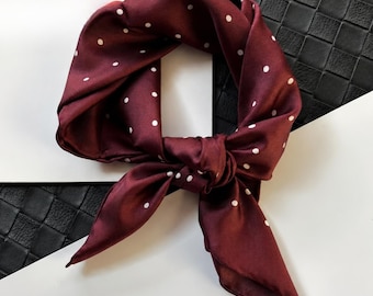 May include: A burgundy silk scarf with a white polka dot pattern, tied in a knot. The scarf is displayed against a black and white background. The scarf's fabric appears smooth and shiny, suggesting a high-quality material. This accessory is suitable for various fashion styles.