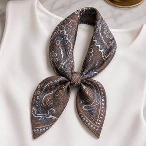 Puede incluir: Un pañuelo cuadrado de color marrón con un patrón de cachemira detallado en tonos azul, blanco y marrón oscuro. El pañuelo está atado en un nudo, mostrando intrincados motivos florales y de lágrima en la tela, sobre una prenda blanca lisa.