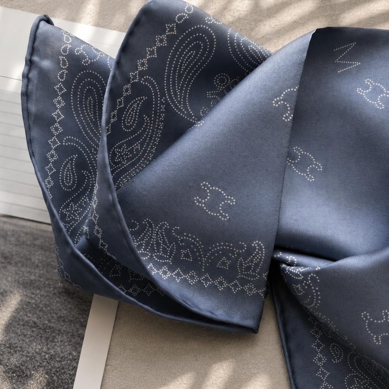 May include: A folded, navy blue silk scarf with a paisley pattern and white dotted accents. The scarf also features a repeating white design. The fabric has a subtle sheen, suggesting a luxurious texture. The scarf is folded in a way that showcases its design.