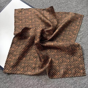May include: A brown pocket square featuring a repeating pattern of small white dots and tiny floral designs. The fabric is folded, displaying its texture and the detailed print. The pocket square is made of a soft, possibly silk, material.