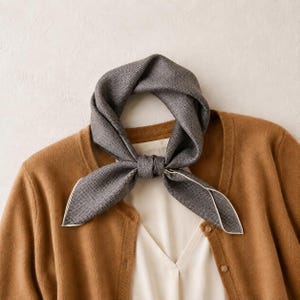 May include: A dark grey square scarf with a fine white dotted pattern and a thin white border, tied in a knot. The scarf is layered over a white top and a tan button-down cardigan, showcasing the textured fabric and the contrast of the colours.