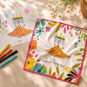 May include: A white silk scarf featuring a colourful floral border and a central illustration of a whimsical figure in a yellow and red striped dress, crowned with a gold tiara. The text "Colors of a Little Soul" is printed on the scarf. Markers and a drawing are also visible.