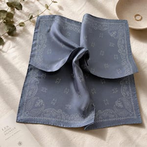 May include: A folded, dusty blue silk scarf with a white paisley and logo pattern. The scarf is square and has a soft, flowing texture. The edges are finished with a delicate white border. The scarf is folded in a way that highlights its design.
