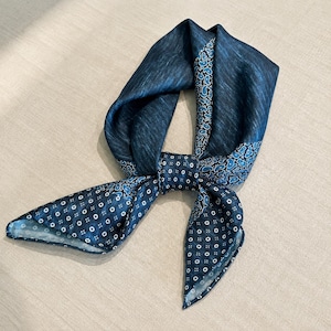 May include: A dark blue neck scarf featuring a mix of patterns including a textured solid blue section, a section with a blue and black paisley print, and a section with a small white geometric square pattern tied in a central knot.