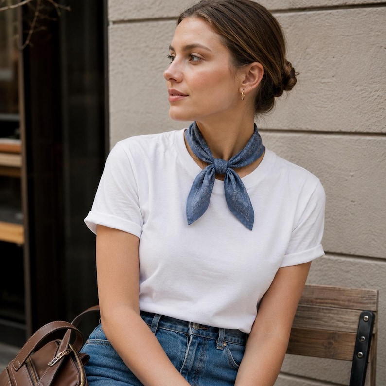 May include: A blue patterned neck scarf tied in a knot. The scarf is worn with a white t-shirt and blue jeans. The person is sitting on a wooden bench.